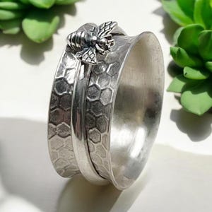 May include: A silver spinning ring with a honeycomb pattern and a bee charm. The ring is made of metal and has a smooth finish.