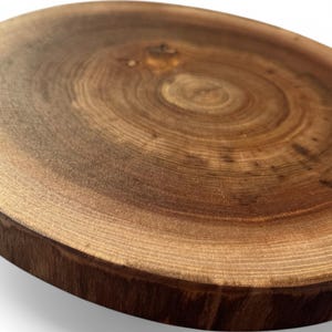May include: A round wooden cutting board with a natural wood grain pattern. The board is made of a dark brown wood with a light brown center.