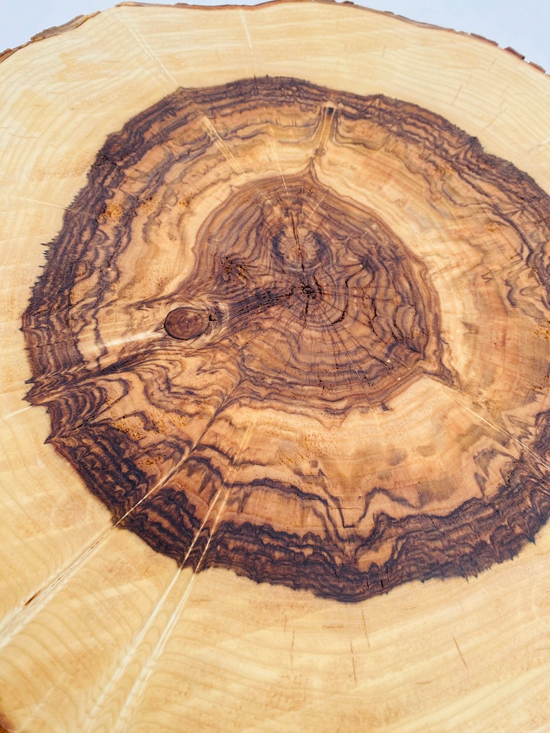 May include: Close-up of a wooden cutting board with a natural wood grain pattern. The board has a circular shape and a dark brown center with lighter brown rings radiating outwards.