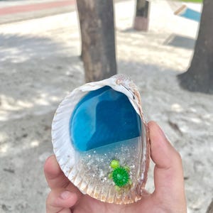 May include: A seashell with a blue resin ocean scene and a small green turtle figurine. The shell is held in a hand, with a sandy beach background.