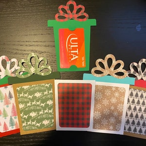 May include: Five Christmas gift card holders made of paper with different patterns. The holders are shaped like presents with a bow on top. The patterns include plaid, snowflakes, Christmas trees, and a green pattern with the words "Very Merry Noel". One holder has a gift card for Ulta Beauty.