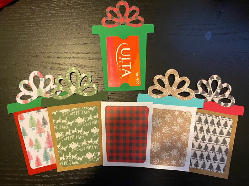 May include: Five Christmas gift card holders made of paper with different patterns. The holders are shaped like presents with a bow on top. The patterns include plaid, snowflakes, Christmas trees, and a green pattern with the words "Very Merry Noel". One holder has a gift card for Ulta Beauty.
