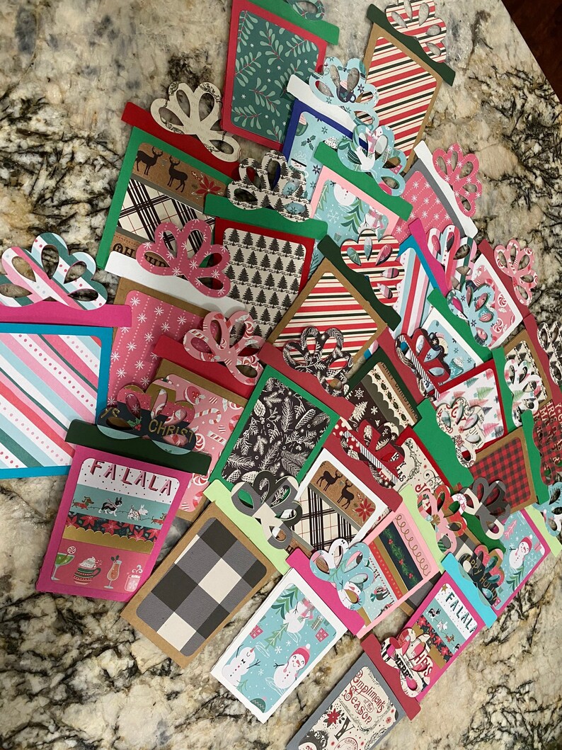 May include: A collection of Christmas-themed gift card holders featuring various patterns and designs, including plaid, snowflakes, and reindeer. The holders are made of paper and have a festive, handmade look.