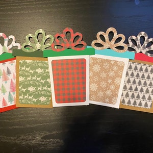 May include: Five Christmas gift card holders with different patterns. The holders are made of paper and have a bow on top. The patterns include a red and black plaid, a brown snowflake pattern, a green reindeer pattern, a green and white Christmas tree pattern, and a gray and white Christmas tree pattern.