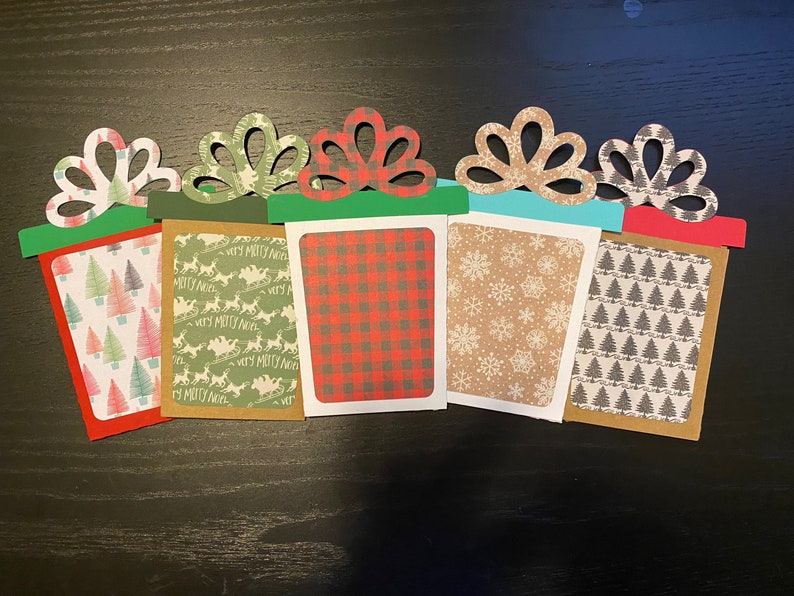 May include: Five Christmas gift card holders with different patterns. The holders are made of paper and have a bow on top. The patterns include a red and black plaid, a brown snowflake pattern, a green reindeer pattern, a green and white Christmas tree pattern, and a gray and white Christmas tree pattern.