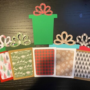 May include: Five Christmas gift boxes with different patterned paper. The boxes are green, red, brown, blue, and white. The boxes have a bow on top. The boxes are on a wooden surface.