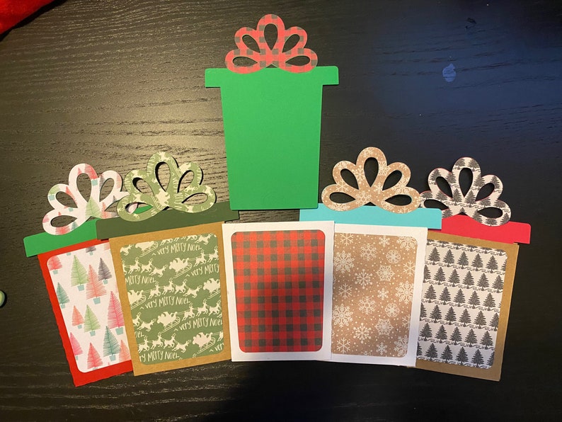 May include: Five Christmas gift boxes with different patterned paper. The boxes are green, red, brown, blue, and white. The boxes have a bow on top. The boxes are on a wooden surface.