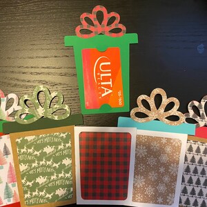 May include: Five Christmas gift card holders with different holiday patterns. The holders are made of paper and have a cut-out design in the shape of a bow. The gift card holders are decorated with various holiday patterns, including snowflakes, Christmas trees, and reindeer. One gift card holder has a green background and a cut-out for a gift card with the text "ULTA BEAUTY" and "25-500".