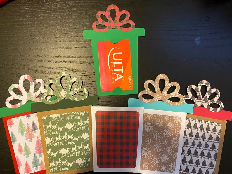 May include: Five Christmas gift card holders with different holiday patterns. The holders are made of paper and have a cut-out design in the shape of a bow. The gift card holders are decorated with various holiday patterns, including snowflakes, Christmas trees, and reindeer. One gift card holder has a green background and a cut-out for a gift card with the text "ULTA BEAUTY" and "25-500".