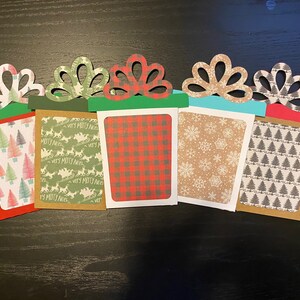 May include: Five Christmas gift card holders with different patterns. The holders are made of paper and have a red, green, brown, and blue color scheme. The patterns include a plaid design, a snowflake design, a Christmas tree design, and a reindeer design.