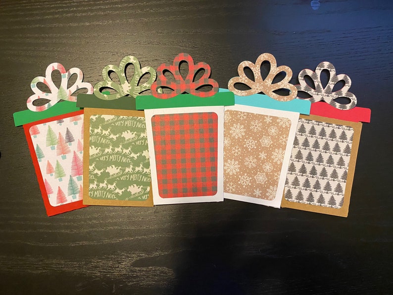 May include: Five Christmas gift card holders with different patterns. The holders are made of paper and have a red, green, brown, and blue color scheme. The patterns include a plaid design, a snowflake design, a Christmas tree design, and a reindeer design.