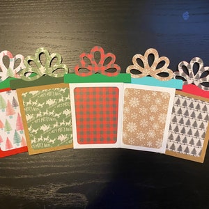 May include: Five Christmas gift card holders with different patterned paper. The holders are decorated with green and red bows and have a brown, red, green, blue, and white background.