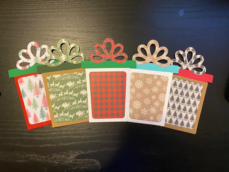 May include: Five Christmas gift card holders with different patterned paper. The holders are decorated with green and red bows and have a brown, red, green, blue, and white background.