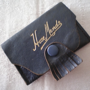 Puede incluir: Una funda de cuero negro con una inscripción dorada que dice "Hose Mends". La funda tiene un botón azul y un detalle de flecos.