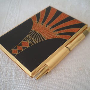 Puede incluir: Un estuche rectangular de metal dorado con un patrón geométrico en forma de abanico negro y naranja en la cubierta frontal. Un bolígrafo dorado a juego está sujeto al lateral del estuche, diseñado para guardar objetos pequeños de forma segura.
