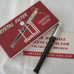 May include: A Dunhill Crystal Filter cigarette holder with a black stem and silver-tone tip, accompanied by its original red rectangular box featuring white text and a graphic illustration, plus an instructional card with red and black text.