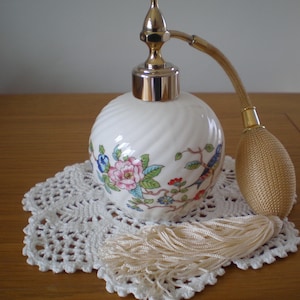 May include: A vintage white perfume bottle with a gold-colored atomizer and a floral design. The bottle sits on a white lace doily, with a matching tassel. The atomizer has a gold-colored bulb.