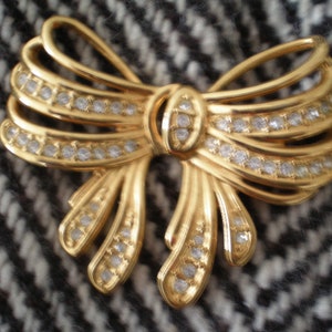 May include: A gold-toned bow brooch with clear rhinestones. The brooch is designed to look like a bow with multiple layers of fabric.