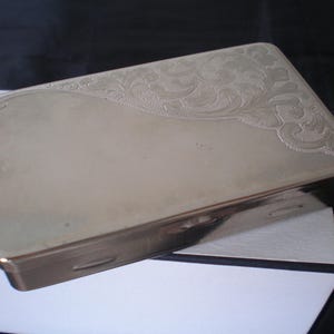 May include: A rectangular silver-colored box with a decorative floral pattern engraved on the top right corner. The box has a smooth, reflective surface and a hinged lid. The box is likely used for storing small items.