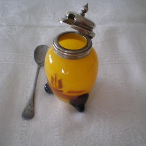 Puede incluir: Un tarro de vidrio amarillo vintage con tapa y borde plateados, y una pequeña cuchara plateada. El tarro tiene una escena pintada de un velero. El tarro descansa sobre tres patas negras.