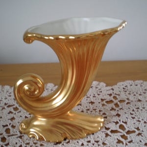 Puede incluir: Un jarrón de cerámica chapado en oro con forma de cuerno estilizado. El jarrón tiene un interior blanco y una base ornamentada y en espiral.