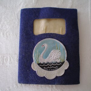 Puede incluir: Un estuche de fieltro azul oscuro con un compacto de agujas vintage. El compacto presenta un emblema plateado con un cisne sobre un fondo azul y verde, con el texto "NEEDLE COMPACT MADE IN ENGLAND". El estuche tiene una ventana rectangular.
