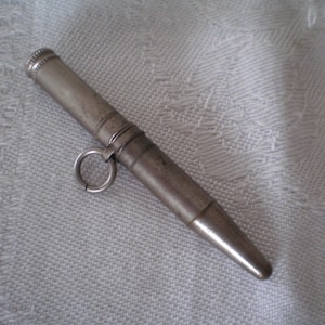 May include: A cylindrical silver-toned metal needle case with a tapered end and a rounded cap. The body features horizontal engraved lines and a small circular ring attached to the side for hanging or attachment purposes, showing a metallic finish.