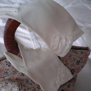 Puede incluir: Un par de guantes blancos lisos hasta la muñeca con un acabado de tela suave y tres pliegues horizontales decorativos en el borde exterior de la muñeca. Los guantes están colocados sobre el asa marrón curvada de un bolso con estampado floral.