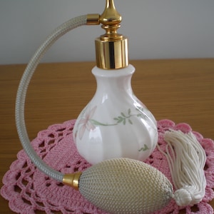 May include: A white porcelain perfume bottle with a floral design and a gold top. The bottle has a long, thin, white tube attached to the top, and a white tassel.