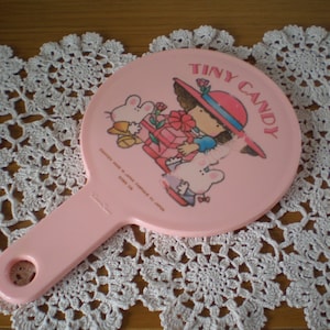 Può includere: Specchio da mano rosa con un'immagine di cartone animato di una ragazza con un cappello rosso e le parole "Tiny Candy".