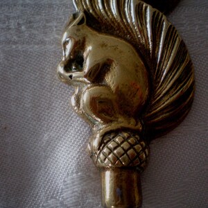 May include: A brass-colored bottle stopper featuring a detailed squirrel design. The squirrel is holding a nut, and the stopper has an acorn-shaped base. The design is highly detailed and has a vintage aesthetic.
