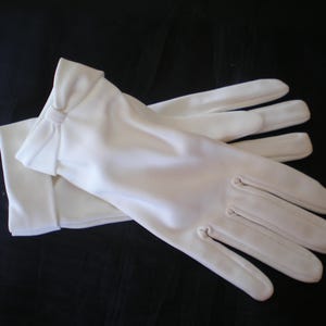 May include: A pair of white gloves, possibly for formal wear, are shown against a dark backdrop. The gloves are made of a smooth material. The design includes a bow detail at the wrist and three stitched lines on the back of each hand.