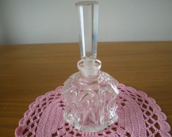 May include: A clear glass bottle with a faceted design and a tall, clear glass stopper. The bottle is sitting on a pink crocheted doily.