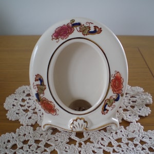 以下が含まれることがあります： 赤、青、緑の色合いの花柄が施された白い陶器製のフォトフレーム。フレームは波打った縁取りとスタンドが付いています。