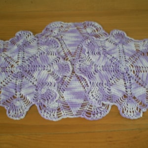 May include: A crocheted table runner in shades of purple and white. The runner has a delicate, lacy design with a scalloped edge.