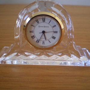 Puede incluir: Un reloj de cristal transparente con un bisel dorado y números romanos. El reloj tiene un diseño facetado y se asienta sobre una base rectangular. La esfera del reloj dice "Genuine Quartz".