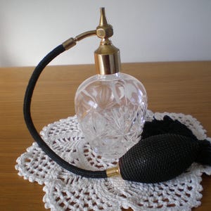 May include: A clear, cut-glass perfume bottle with a gold-colored atomizer and black bulb. The bottle sits on a white, crocheted doily. The atomizer has a curved black tube. The bottle is round with a gold-colored top.