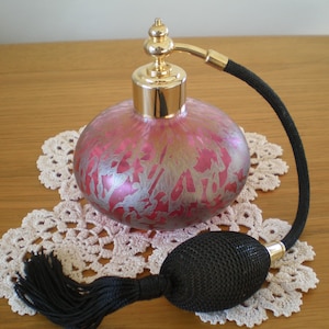May include: A vintage perfume bottle with a pink and silver design, gold accents, and a black atomizer bulb. The bottle sits on a white lace doily, showcasing a classic, elegant aesthetic.