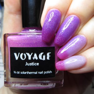 Puede incluir: Un frasco de esmalte de uñas solarthermal "VOYAGE Justice" con tapa negra. El esmalte de uñas es de color púrpura brillante, y las uñas están pintadas con un color a juego que se desvanece de púrpura a rosa.