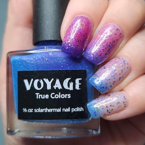 以下が含まれることがあります： 「VOYAGE True Colors」の文字が入った青色のソーラーサーマルネイルポリッシュのボトル。爪はピンク、紫、青のグラデーションで、グリッターが施されています。ボトルには1/2オンスのポリッシュが入っています。