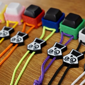 May include: Nine colorful keychains with black and white patterned charms. Each keychain features a small square keycap with a different colored fabric covering. The keycaps are attached to a cord with a black and white charm.