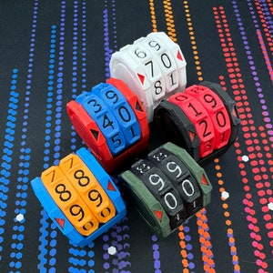以下が含まれることがあります： カラフルな3Dプリントされた数字ホイールのコレクション。ホイールは白、赤、青、オレンジ、黒など、さまざまな色があります。各ホイールには一連の数字が表示され、現在の数字を示す赤い三角形があります。ホイールはパターン化された表面に配置されています。