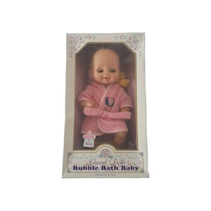 May include: A vintage Laurel Dolls Bubble Bath Baby doll in its original packaging. The doll is wearing a pink robe with a matching belt and a small graphic. The box features the text "Bubble Bath Baby" and "Morning Eyes All Vinyl".