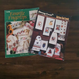 Può includere: Due libri di artigianato su una superficie di legno. Il libro verde, "Favorite Fingertips", presenta disegni floreali e di frutta. L'altro, "Stoney Creek Collection", mostra modelli di asciugamani da cucina ricamati con vari motivi, tra cui galli e frutta.