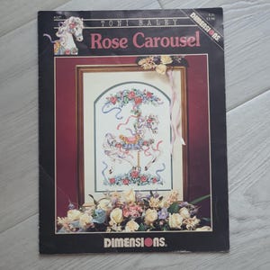 May include: A cross-stitch kit titled "Rose Carousel" by Toni Baley. The kit features a carousel horse design with roses and ribbons. The kit's cover is black with a floral arrangement at the bottom and a carousel horse illustration at the top.