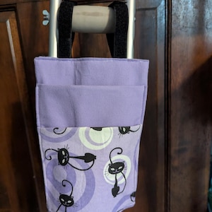 May include: A lavender fabric pouch with two black hook-and-loop straps attached to the top. The lower section features a pattern of black cats on a background of light purple and white circles. The pouch is suspended from a metal crutch handle.