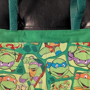 May include: A green fabric tote bag featuring a repeating pattern of Teenage Mutant Ninja Turtles characters in various poses, including those wearing blue, purple, orange, and red masks, with green fabric straps attached to a black leather-like top section.