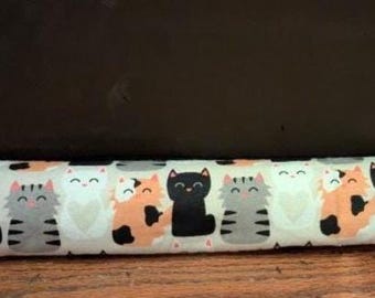 Può includere: Un paraspifferi cilindrico e lungo in tessuto con un motivo ripetuto di gatti in stile cartoon di vari colori, tra cui nero, grigio, bianco e calicò arancione, tutti con semplici volti sorridenti su uno sfondo chiaro.