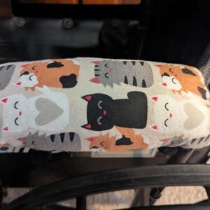 May include: A rectangular armrest cover featuring a repeating pattern of cartoon cats in black, white, gray, and orange on a light beige background. The fabric cover is secured over a black armrest, displaying various feline expressions.