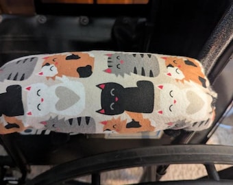 May include: A rectangular armrest cover featuring a repeating pattern of cartoon cats in black, white, gray, and orange on a light beige background. The fabric cover is secured over a black armrest, displaying various feline expressions.